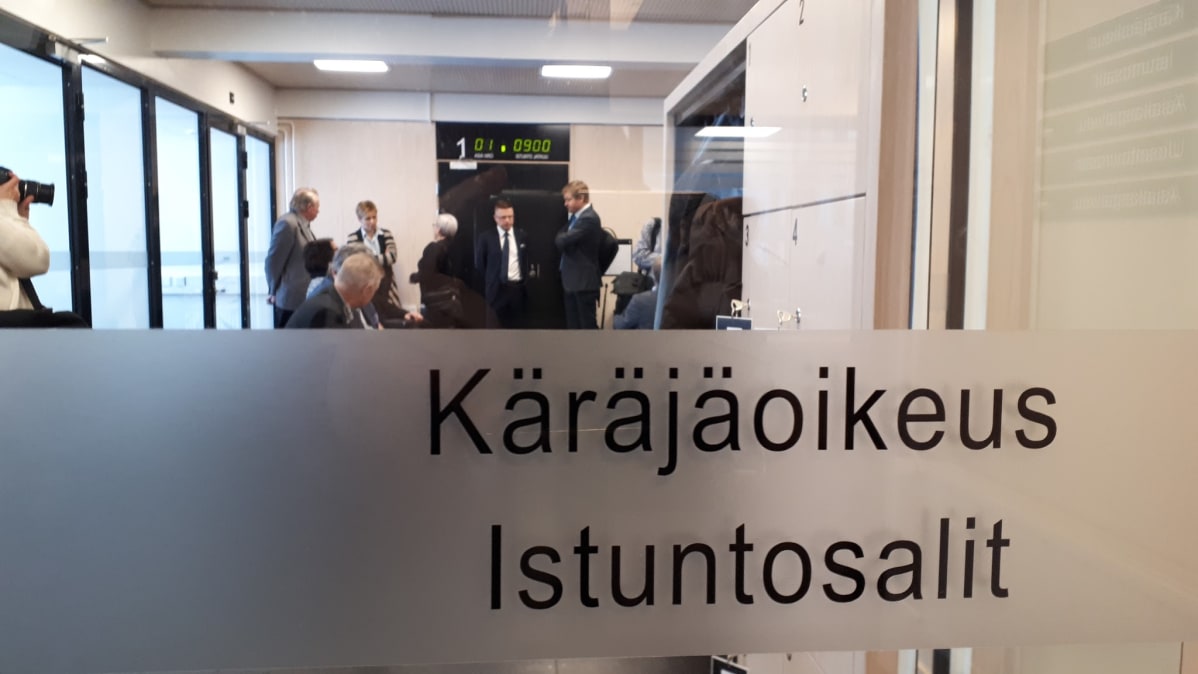 Ylivieska-Raahen käräjäoikeuden istustosalin lasiovi, jonka takana henkilöitä odottamassa pääkäsittelyn alkua.