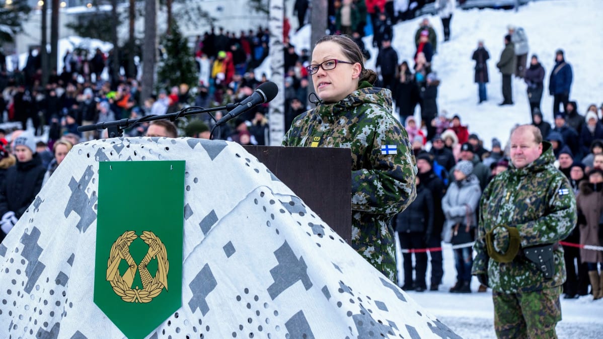 Sanna Timonen puhujanpöntön takana puhumassa mikrofoniin armeijauniformussa. Taustalla seisoo paljon ihmisiä kuuntelemassa.
