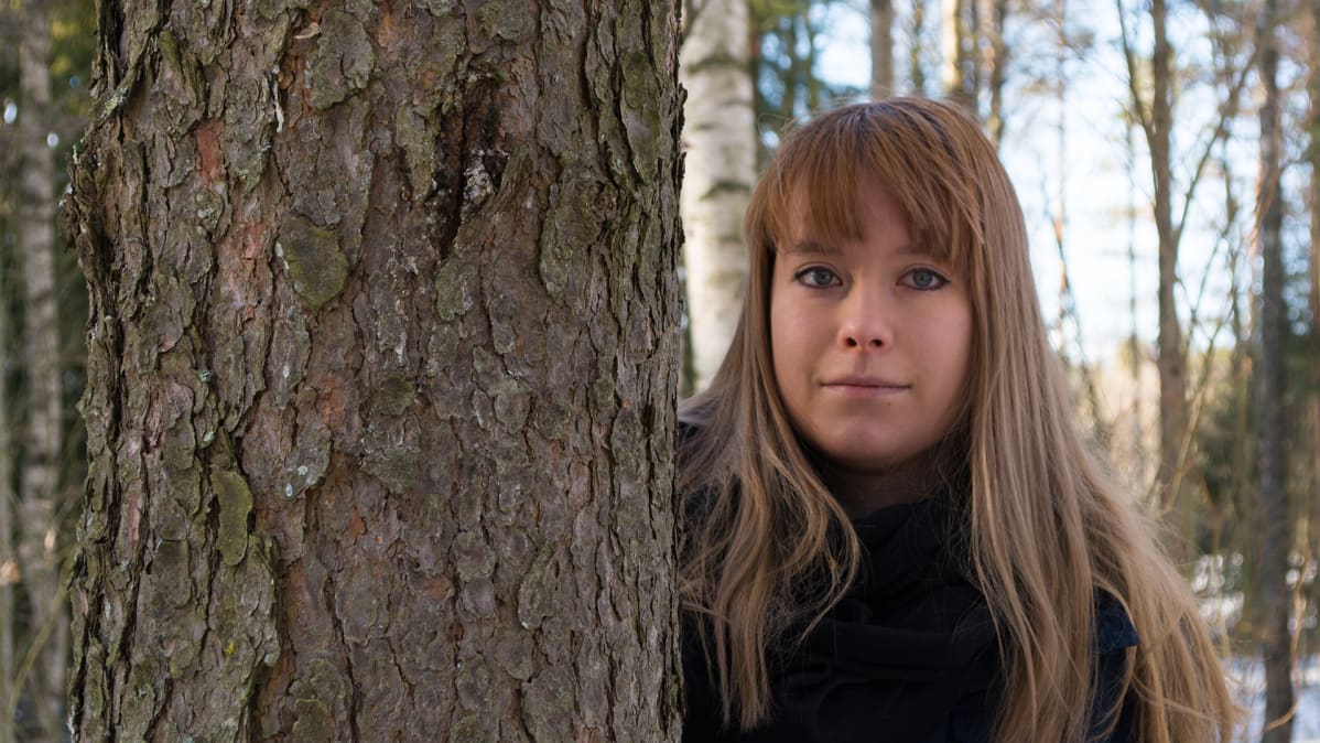 Vaalea nainen katsoo männyn rungon takaa