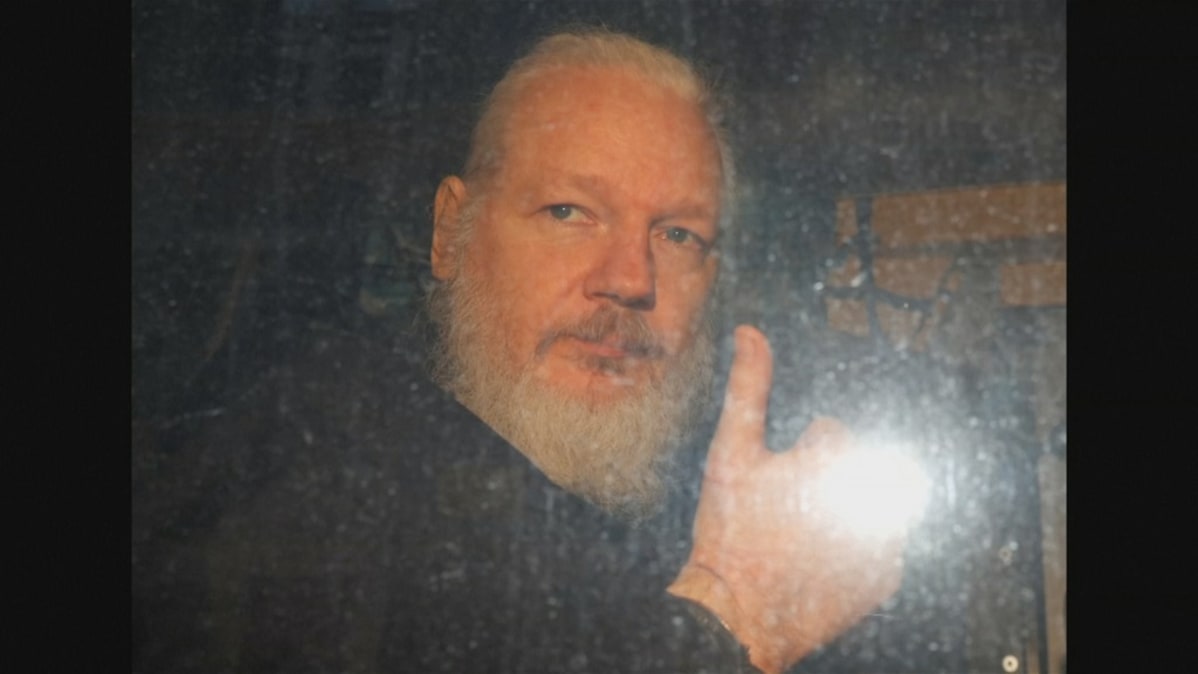 Kuvassa Julian Assange poliisiauton kyydissä. Hän näyttää oikealla kädellään peukaloa.