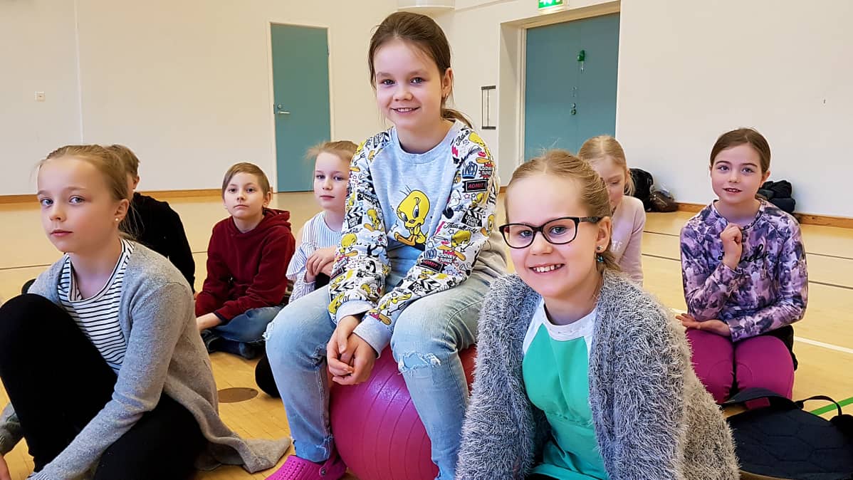 lapsia koulun liikuntasalin lattialla, oopperaharjoitukset Haukkavuoren koululla Kotkassa, Olga Metelgina pallon päällä , vieressä oikealla Elina Kamppinen