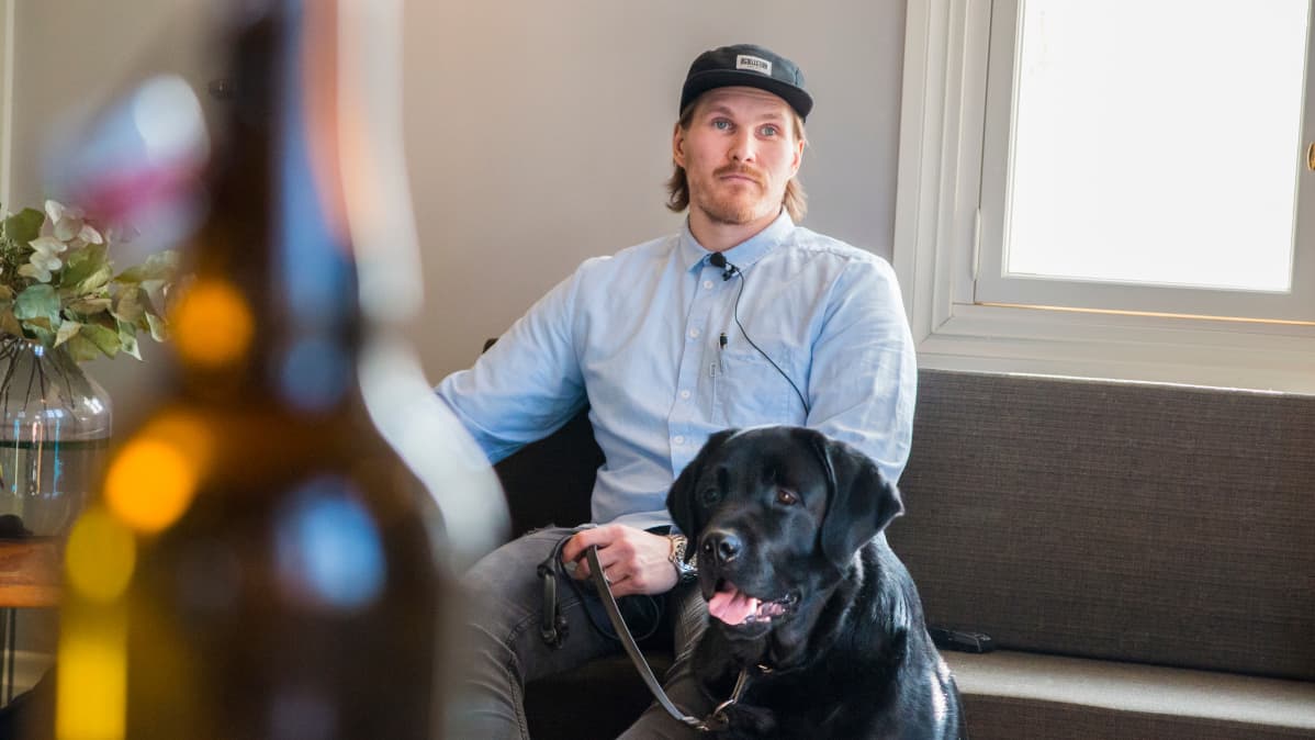 Mies istuu sohvalla vieressään musta labradorinnoutaja.