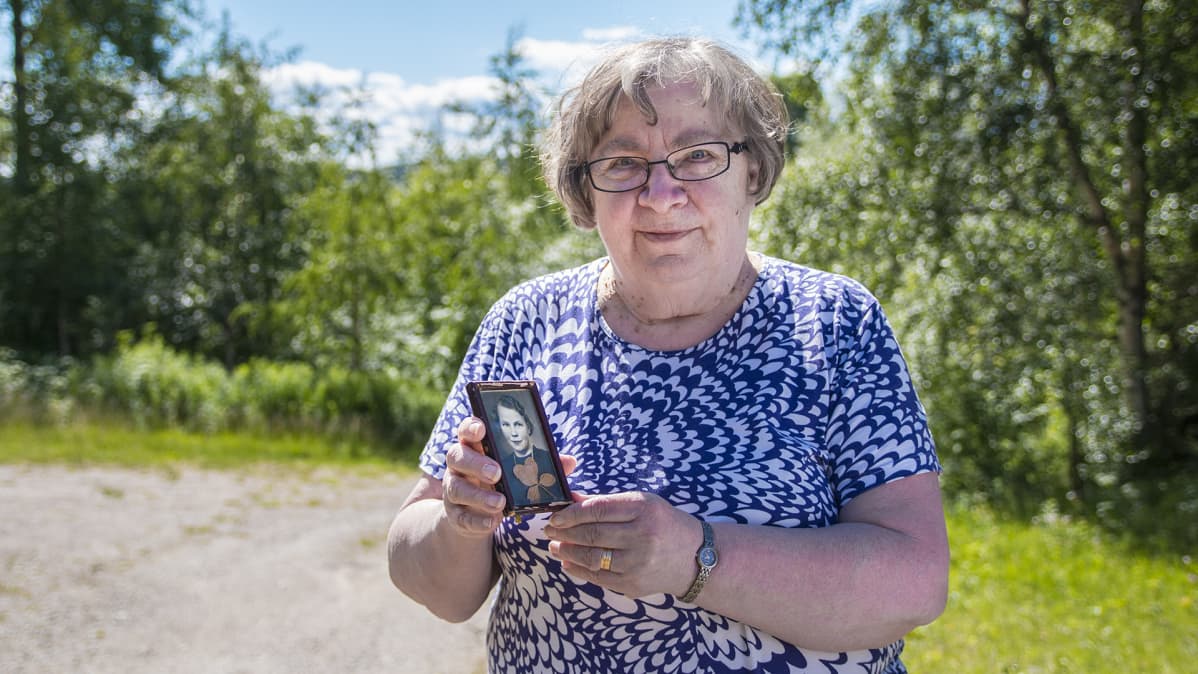 Raili Kemppainen seisoo ulkona auringon paisteessa ja pitelee kuvaa äidistään Ellistä.