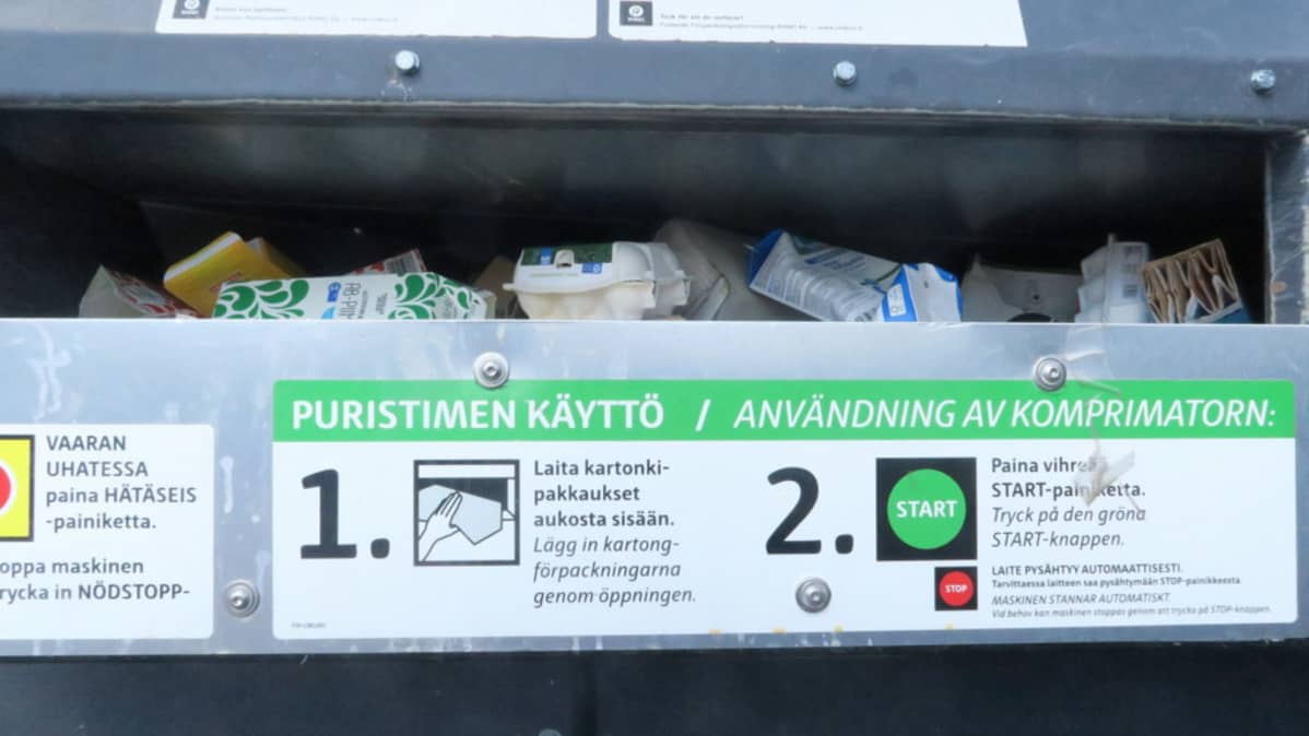 Kartonkikeräyspuristimen täyttöaukossa pakkauksia, alla puristimen käyttöohje