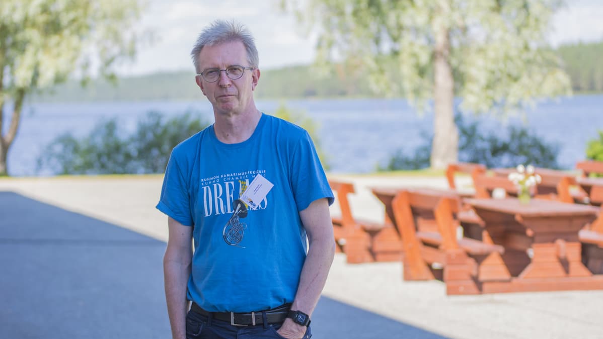 Mies seisoo ulkona. Takana on puupöytiä ja järvimaisema.