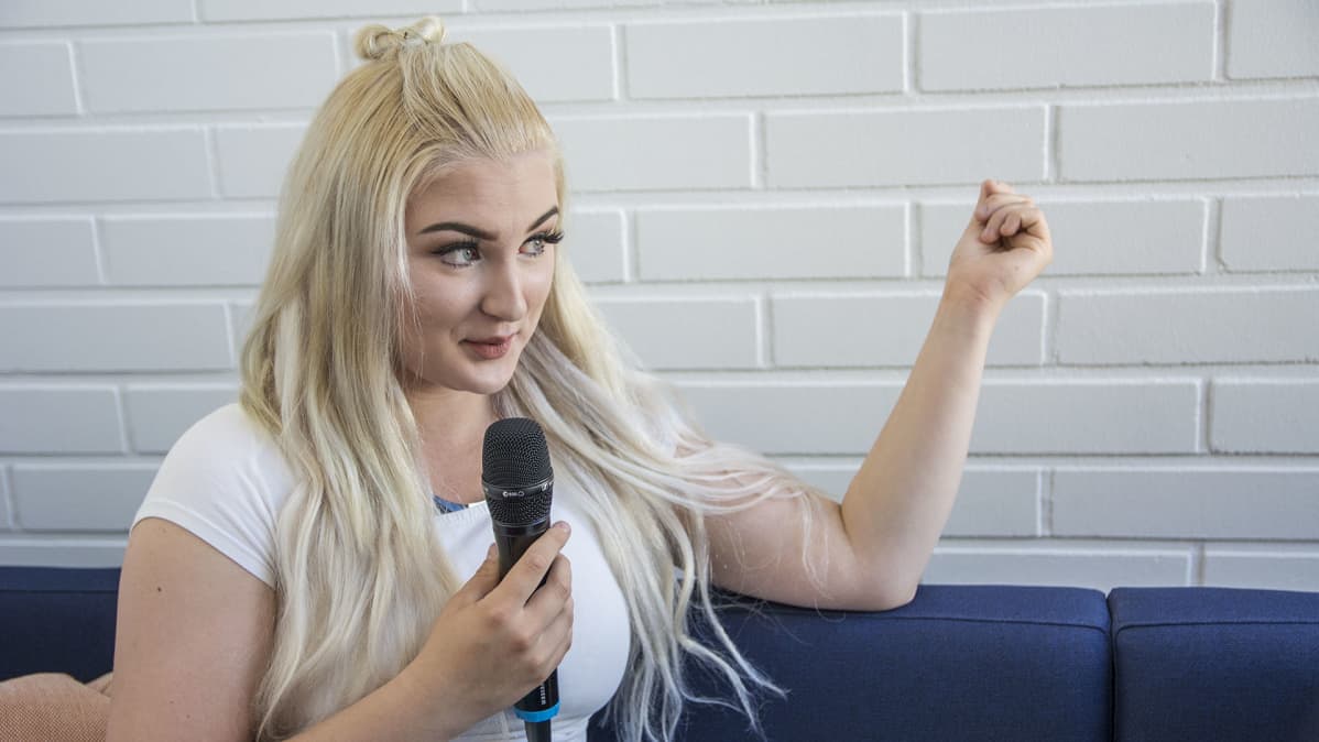 Nainen juttelee mikki kädessä ja istuu sohvalla.