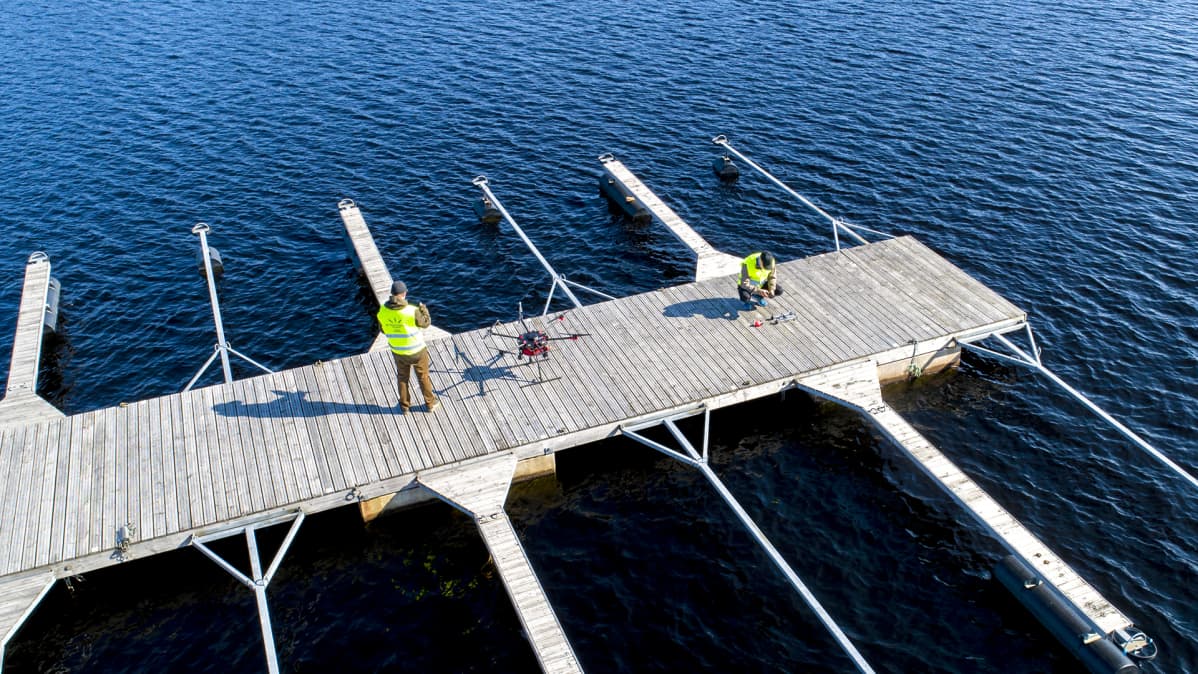 Ilmakuvassa kaksi miestä laittaa suurta dronea lähtökuntoon venelaiturilla.