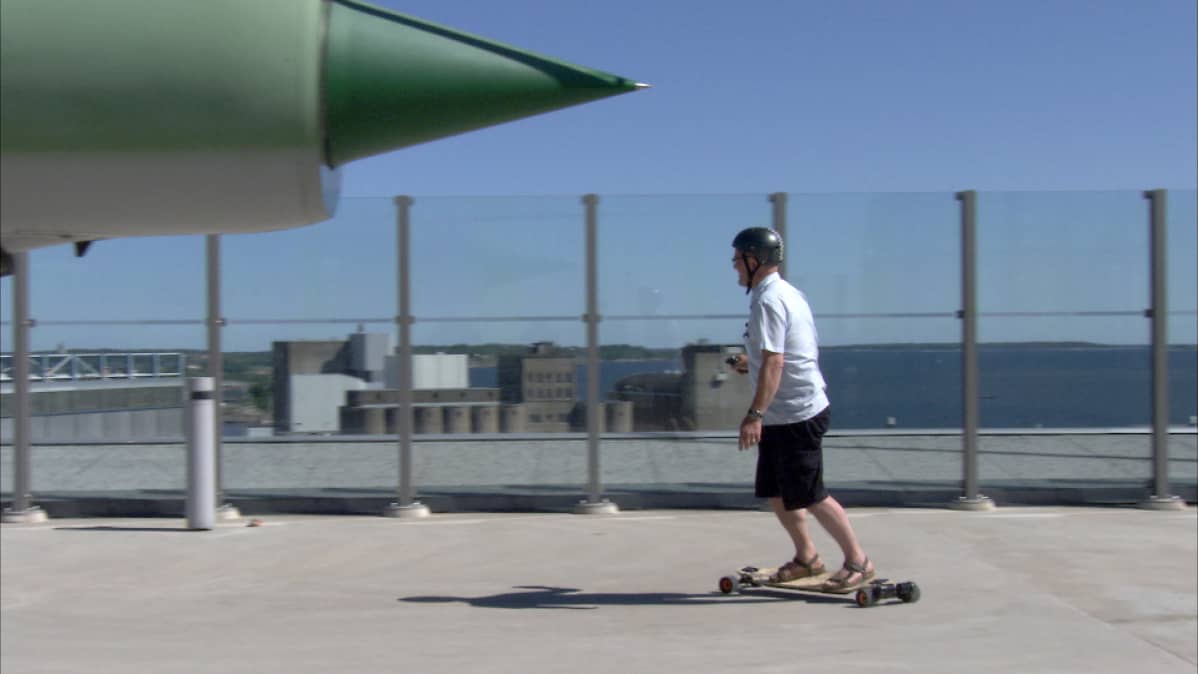 Evolve Bamboo GT Street sähkölongboard Verkkokaupan kattoterassilla Jätkäsaaressa. Toimittaja Matti Toivonen kokeilee (kypärä päässä).