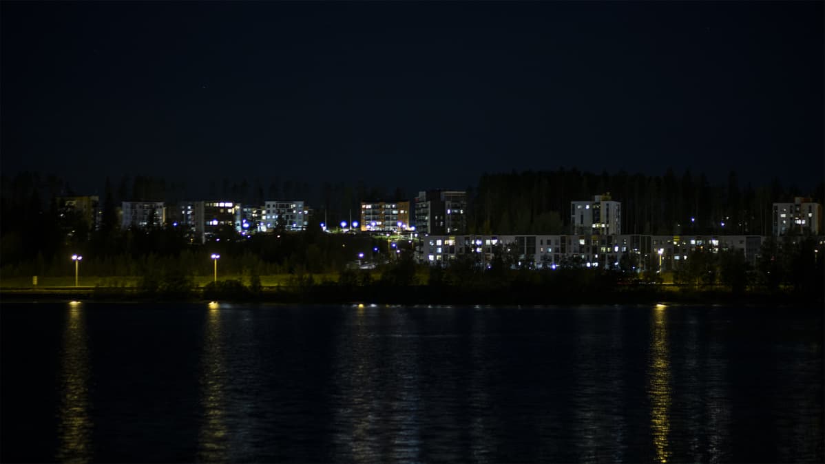Pimeä kaupunki, valaistuja taloja järven takaa kuvattuna.