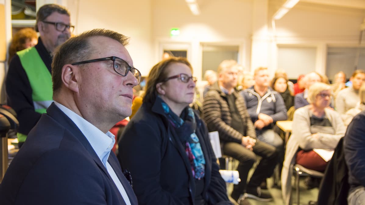 Yleisötilaisuus, etualalla silmälasipäinen mies katsoo oikealle tarkkaavaisena.