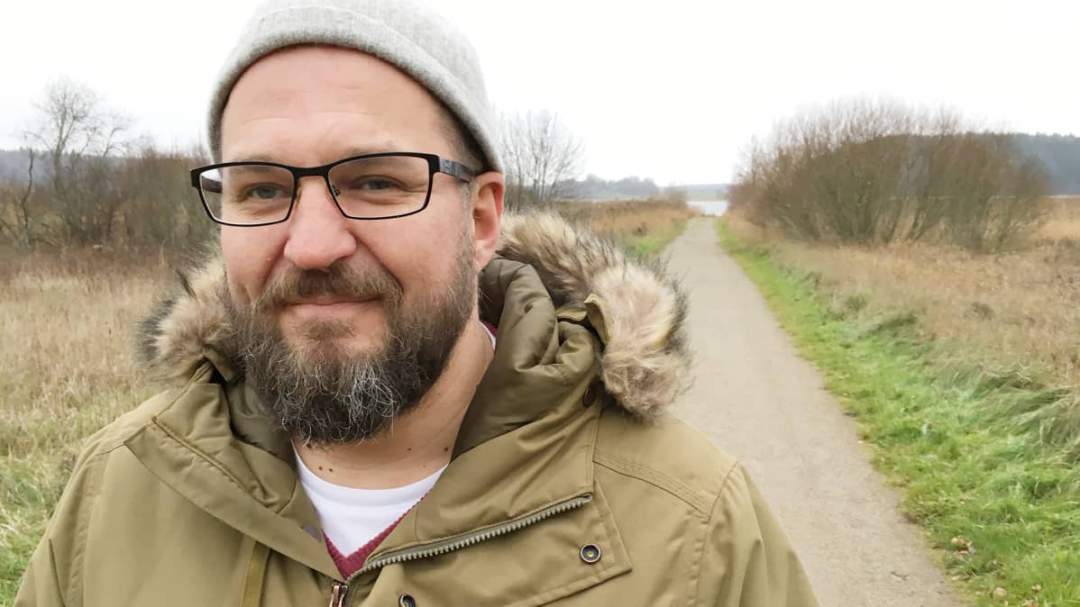 Parrakas, silmälasipäinen mies talvitakissa ja pipo päässä järjenranta taustalla.