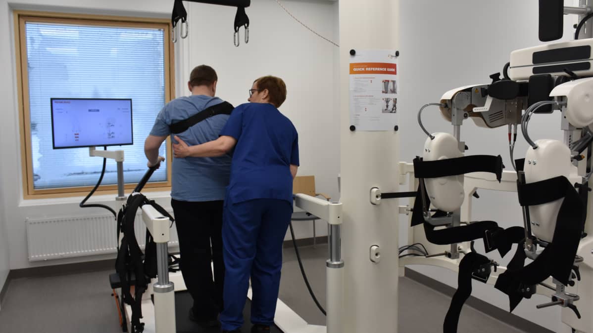Kuvassa hoitaja pukee kuntoutujalle kävelyrobotin valjaita