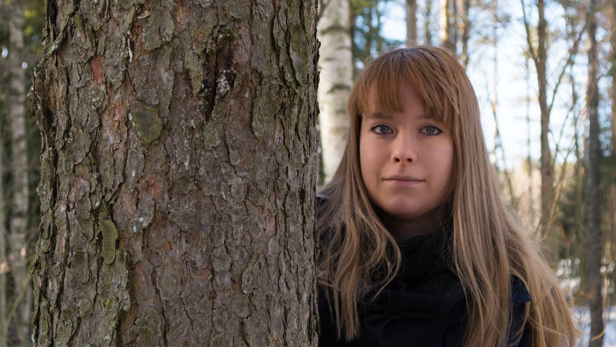 Vaalea nainen katsoo männyn rungon takaa