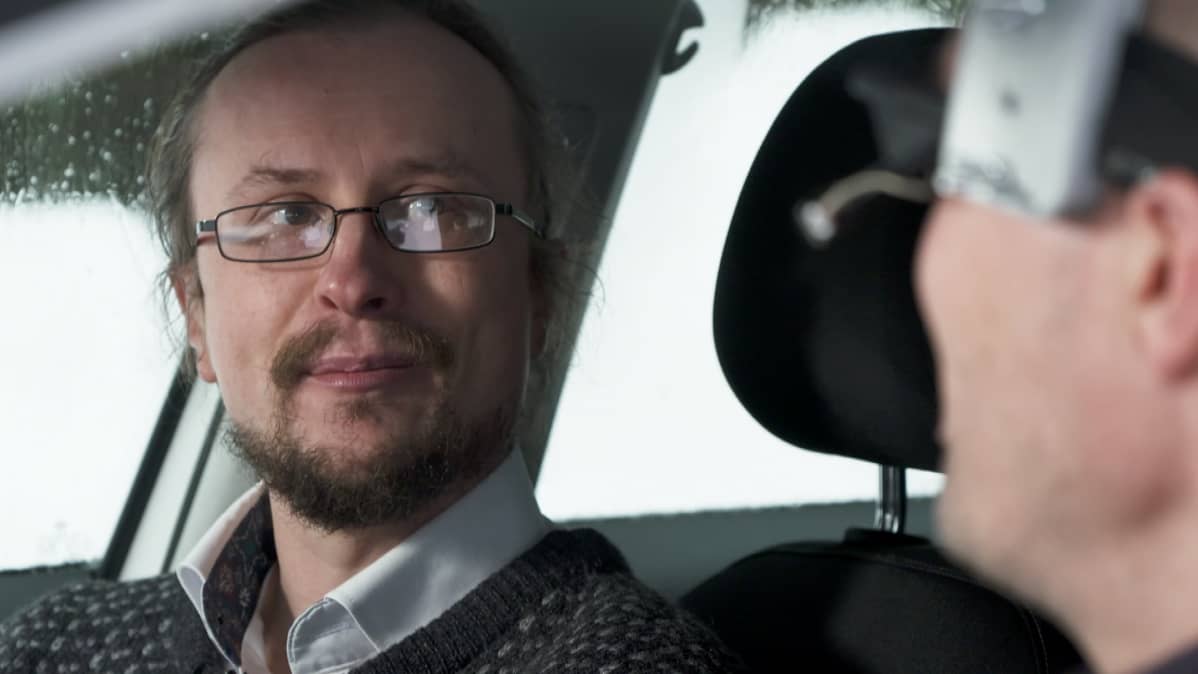 Silmälasipäinen mies istuu autossa.