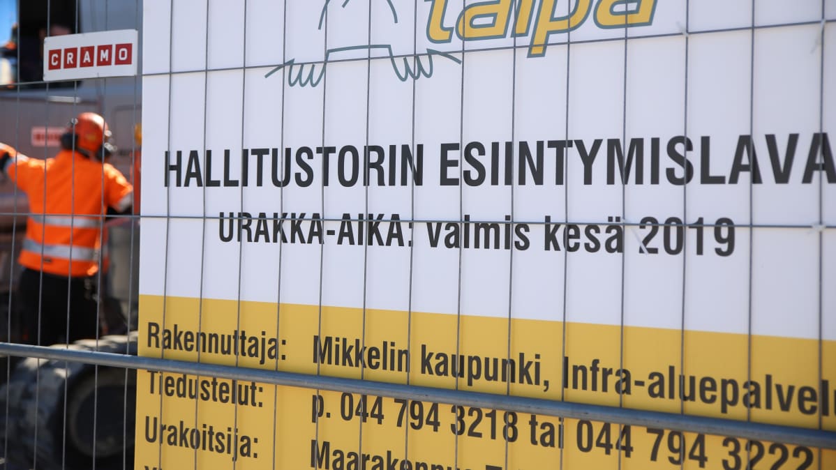 Kuva kyltistä, jossa kerrotaan esiintymislavan rakentamisesta. Taustalla rakennustyömies oranssissa takissa.