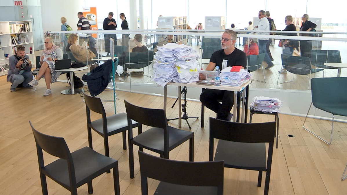 Kansanedustaja (Kok.) ja dekkarikirjailija Marko Kilpi lukee sote-lakiesitystä keskustakirjasto Oodissa 1.8.2019 Puoli seitsemän -ohjelman Lukuhaaste-tempauksessa.. Paperipinoja pöydällä, viisi tyhjää tuolia lukijan edessä.