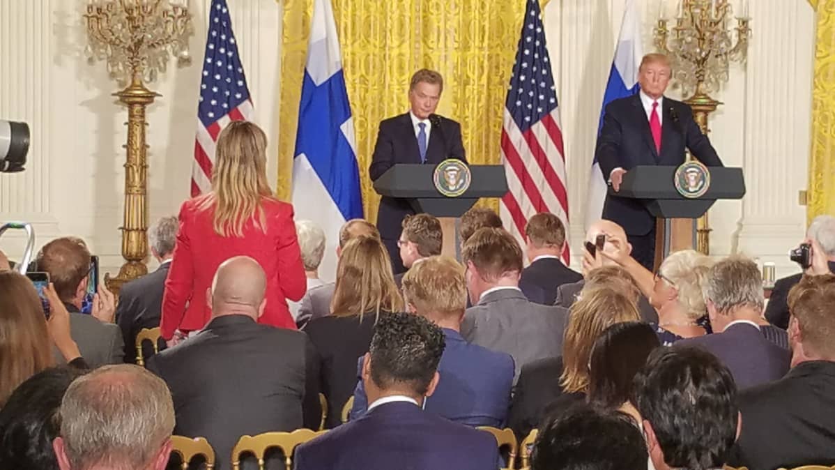 Kuvassa etualalla toimittaja punaisessa takissa väkijoukon keskellä. Taka-alalla presidentti Sauli Niinistö ja presidentti Donald Trump.