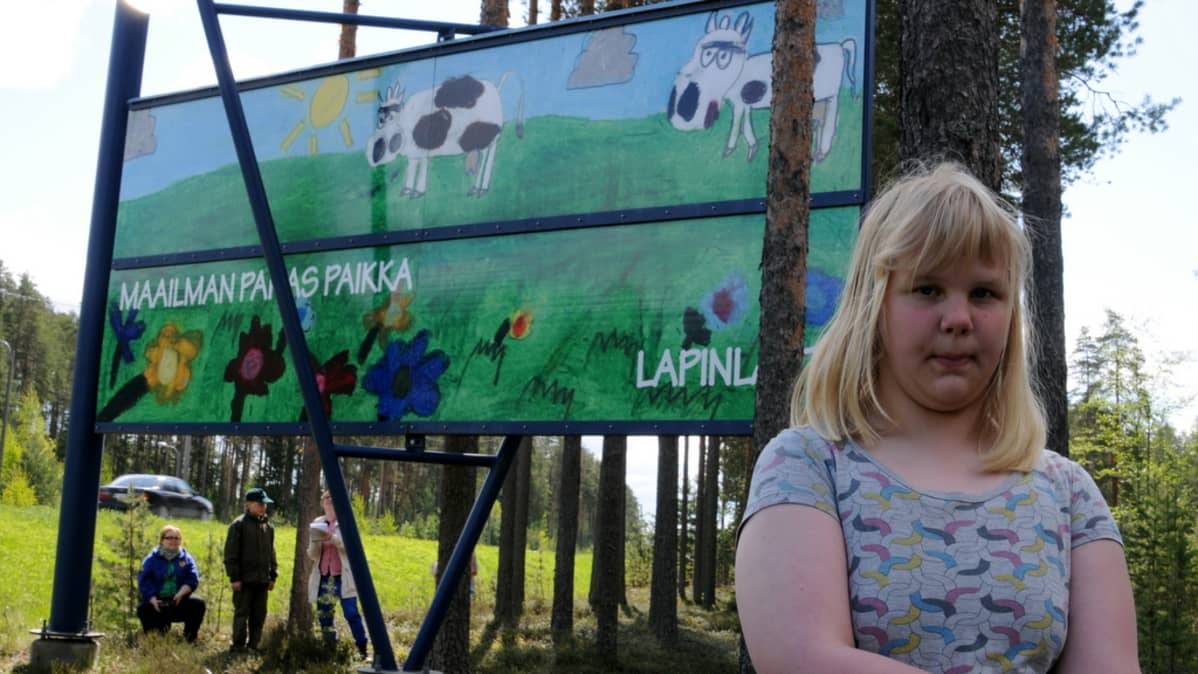 Karoliina Huttunen seisoo kuvittamansa tienvarsitaulun edessä.