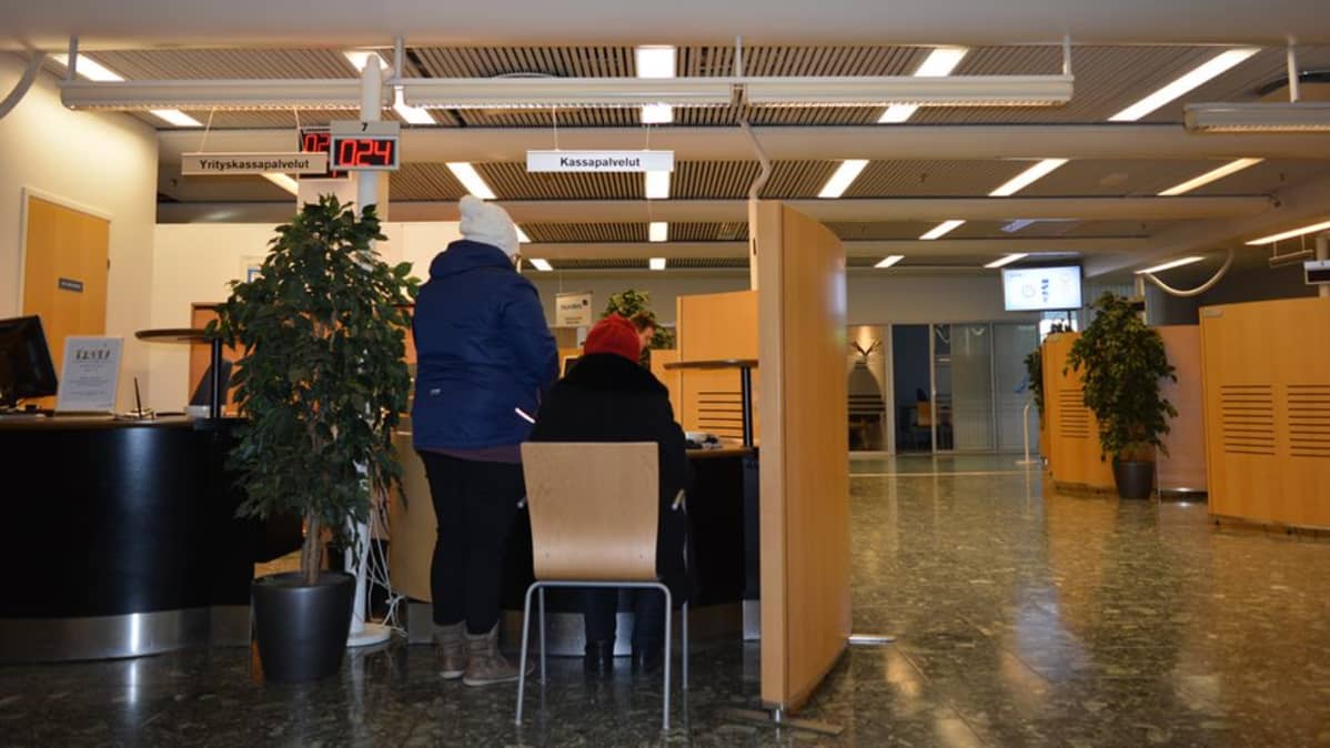 Kaksi asiakasta pankin kassapalveluiden asiakaspisteellä.