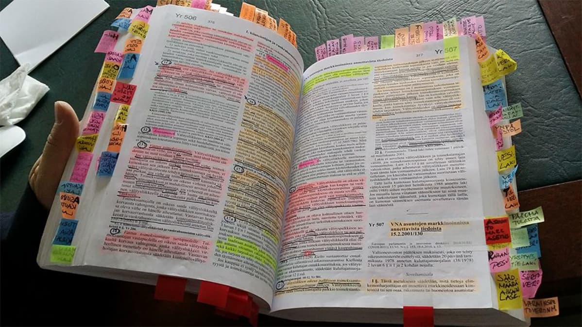 Kuvassa avoin lakikirja, jossa on viihattuja ja korostettuja tekstejä, värikkäitä postit-lappuja