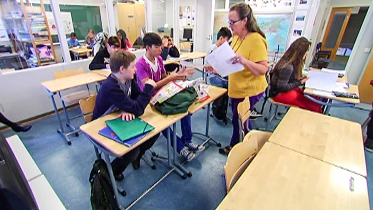 opettaja luokassa pulpeteissa istuvien oppilaiden keskellä