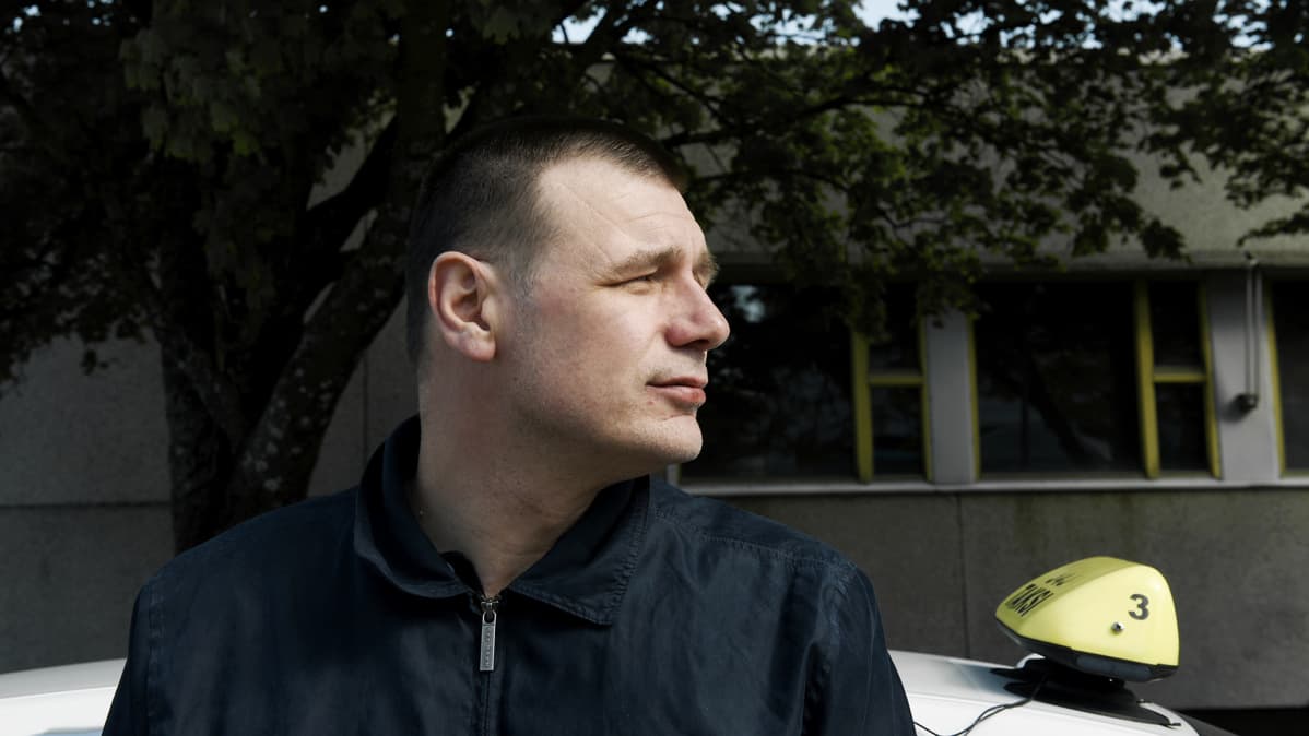 Tummaan asuun pukeutunut Vilho Sakari Leino nojaa valkoiseen taksiautoon.