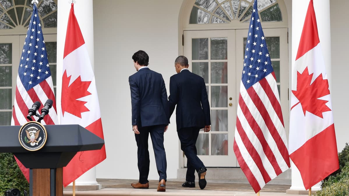 Justin Trudeau ja Barack Obama kävelevät selin kameraan Valkoisen talon kuistille suurten Yhdysvaltain ja Kanadan lippujen välistä.