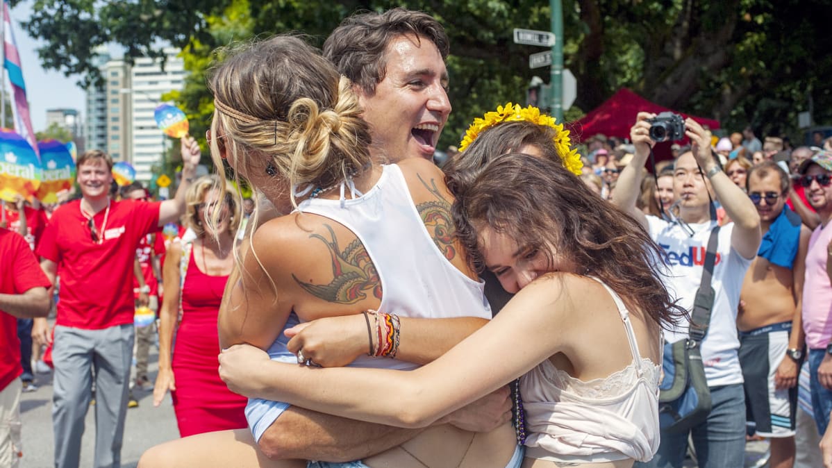 Kolme naista halaa Justin Trudeauta, taustalla ihmiset ottavat tilanteesta kuvia.