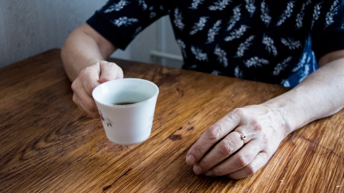 Henkilö istuu pöydän ääressä ja pitelee toisessa kädessä kahvikuppia.
