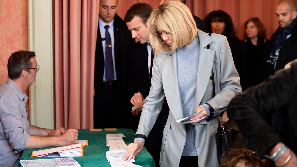 Brigitte Trogneux ja Emmanuel Macron poimivat äänestyksessä tarvittavia lappusia vaalivirkailijan valvomalta pöydältä. Trogneux'lla on vaaleanharmaa päällystakki. Macronilla on tumma puku.