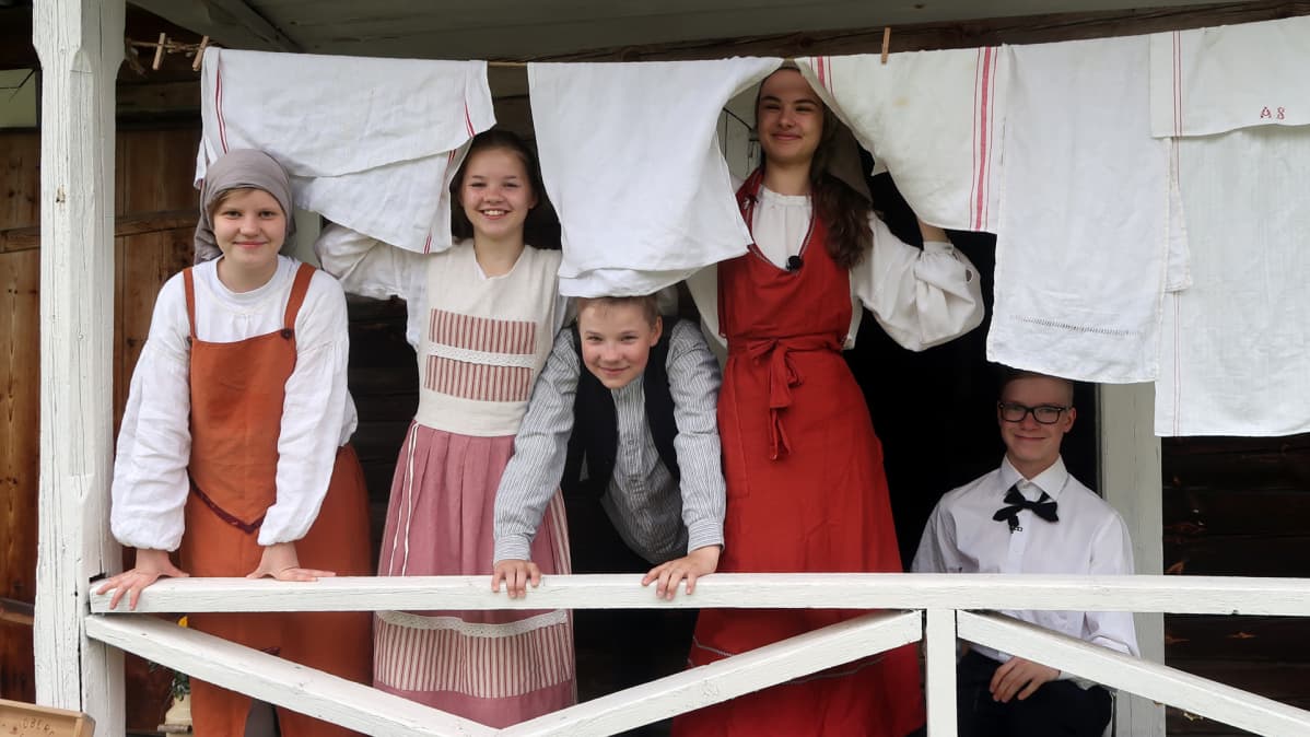 Kuvassa nuoria 1800-luvun vaatteissa pyykinarun takana.