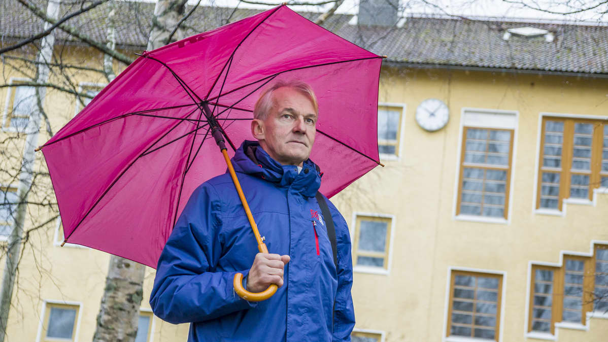 Mies seisoo sateessa sateenvarjon alla vanhan rakennuksen edessä.