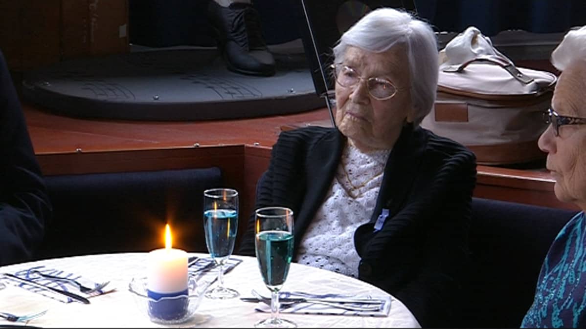 Lottaveteraani istuu ravintolan pöydässä veteraanilounaan alkaessa., pöydällä kybnttilä ja tervetuliaismalja