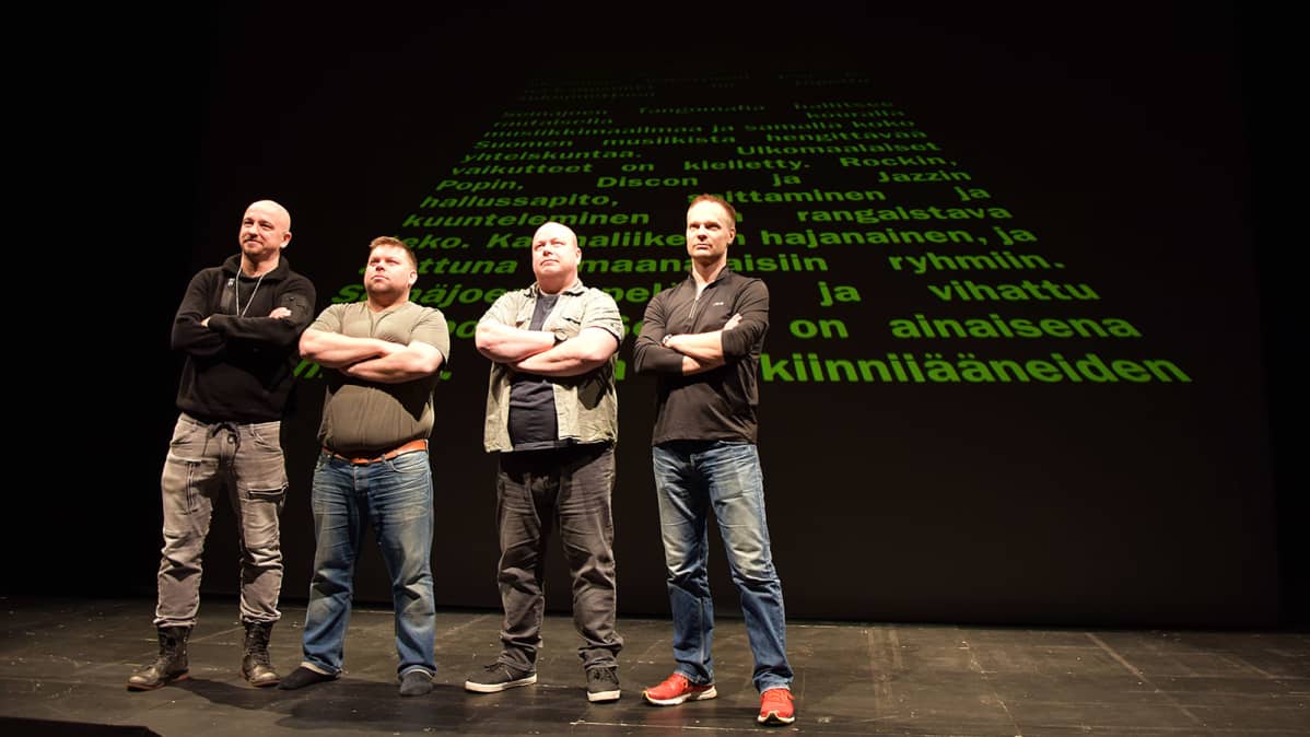 Neljä miestä seisoo teatterin lavalla kädet puuskassa, taustalla Tähtien Sodasta lainattu alkutekstiteema