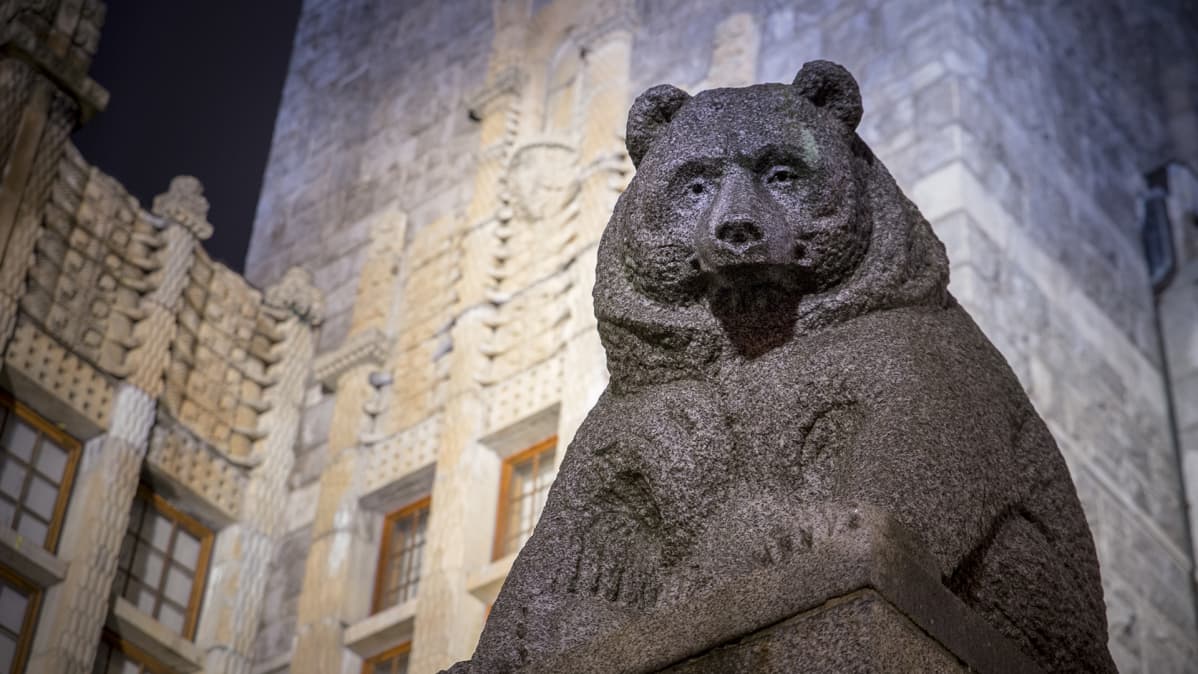 Suomen kansallismuseo iltavalaistuksessa, kuvassa karhu-patsas etualalla.