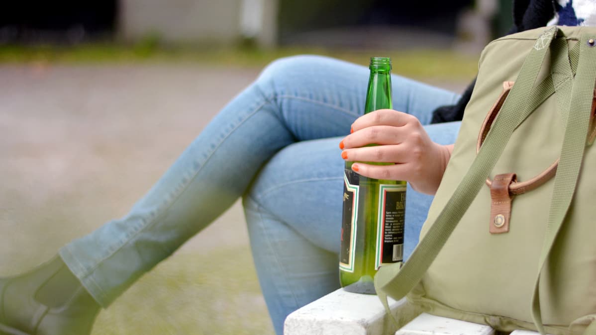 En person sitter på en bänk med en ryggsäck och vinflaska bredvid sig.
