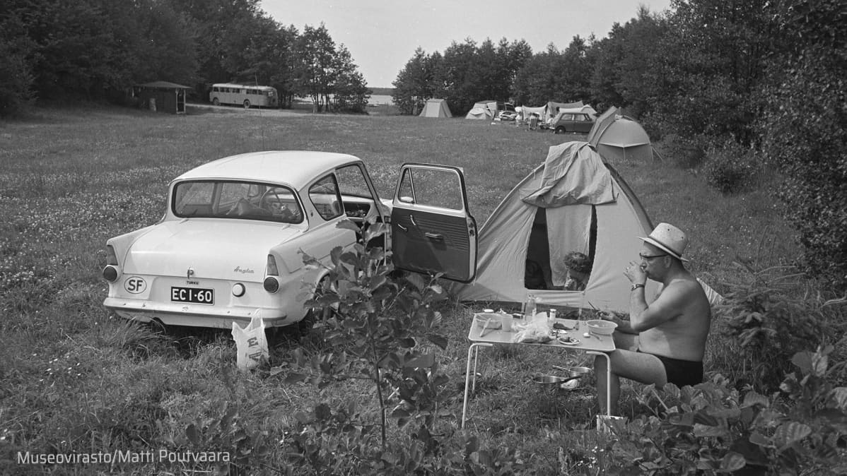 Ford Anglia leirintäalueella, kupoliteltan edessä mies retkipöydän ääressä uimahousuisillaan.