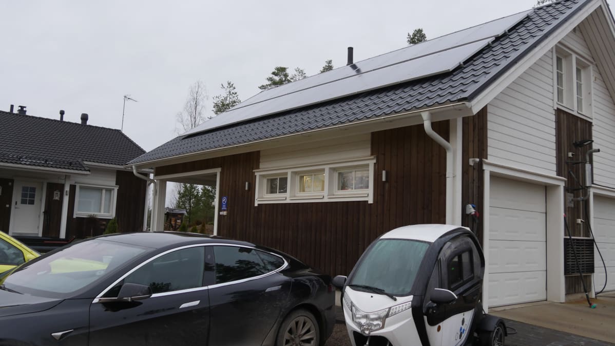 Autotallin katolla aurinkopaneelit. Pihassa sähköauto ja sähkömopo.