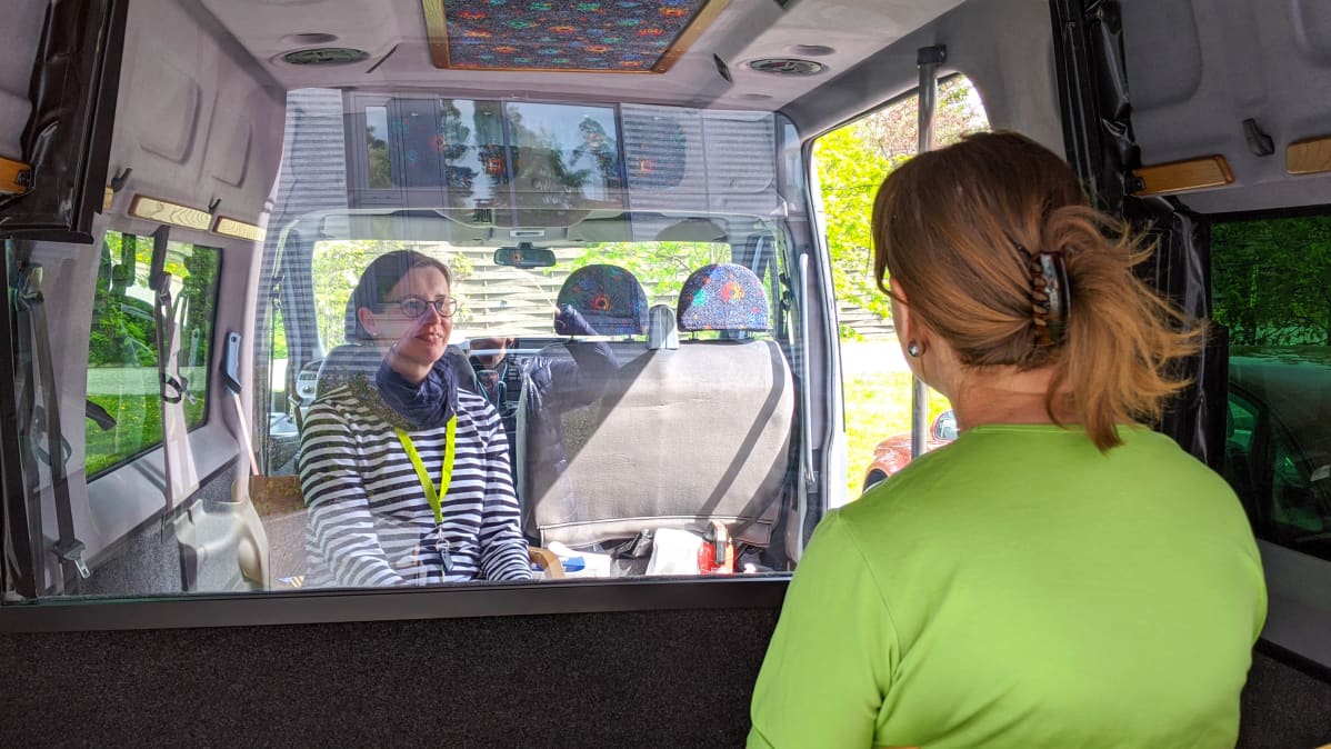 Simonkylän hoivakodin asumispalvelujen esimies Sanna Kostamo istuu vanhusten tapaamisbussissa pleksin takana.