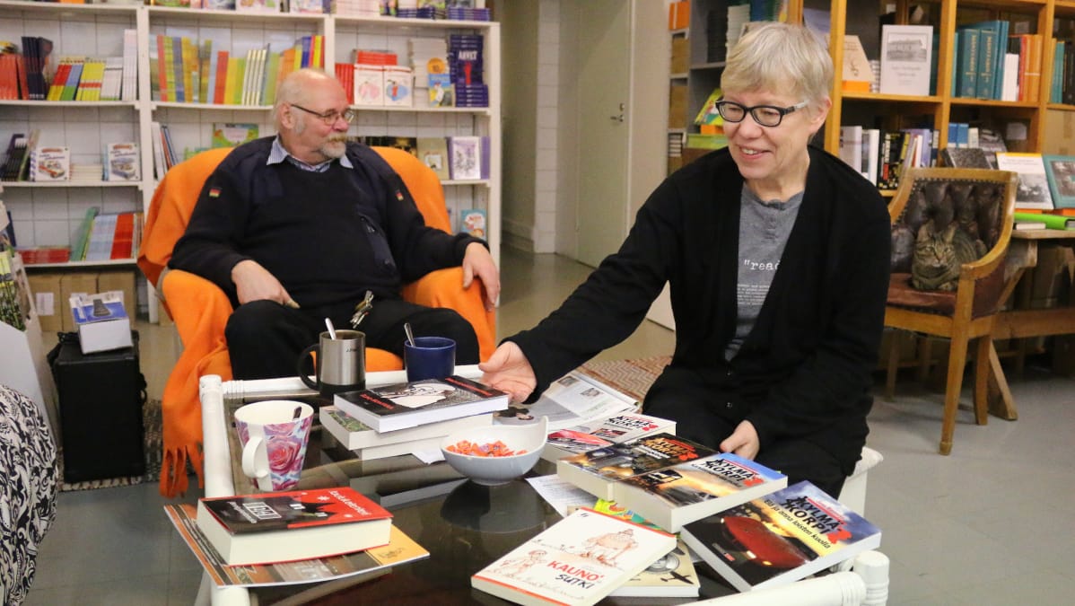 mies ja nainen istuvat pöydän ääressä, pöydällä kirjoja ja kahvikuppeja, taustalla kissa nojatuolissa