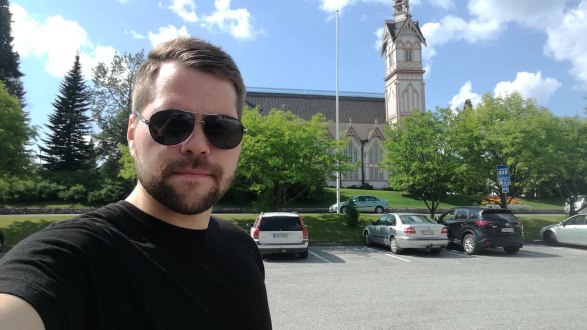 Nuori mies ottaa selfien parkkipaikalla. Taka-alalla on Kajaanin kirkko.