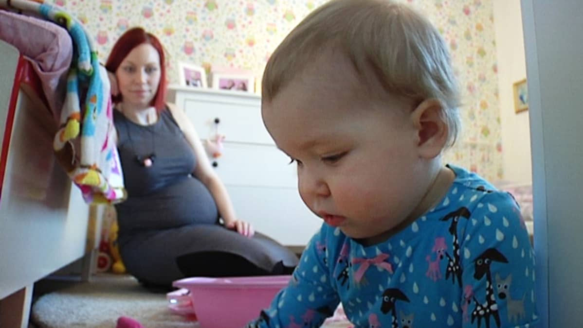 1,5-vuotias lapsi huoneessaan, äiti seuraa taustalla lapsen leikkimistä.
