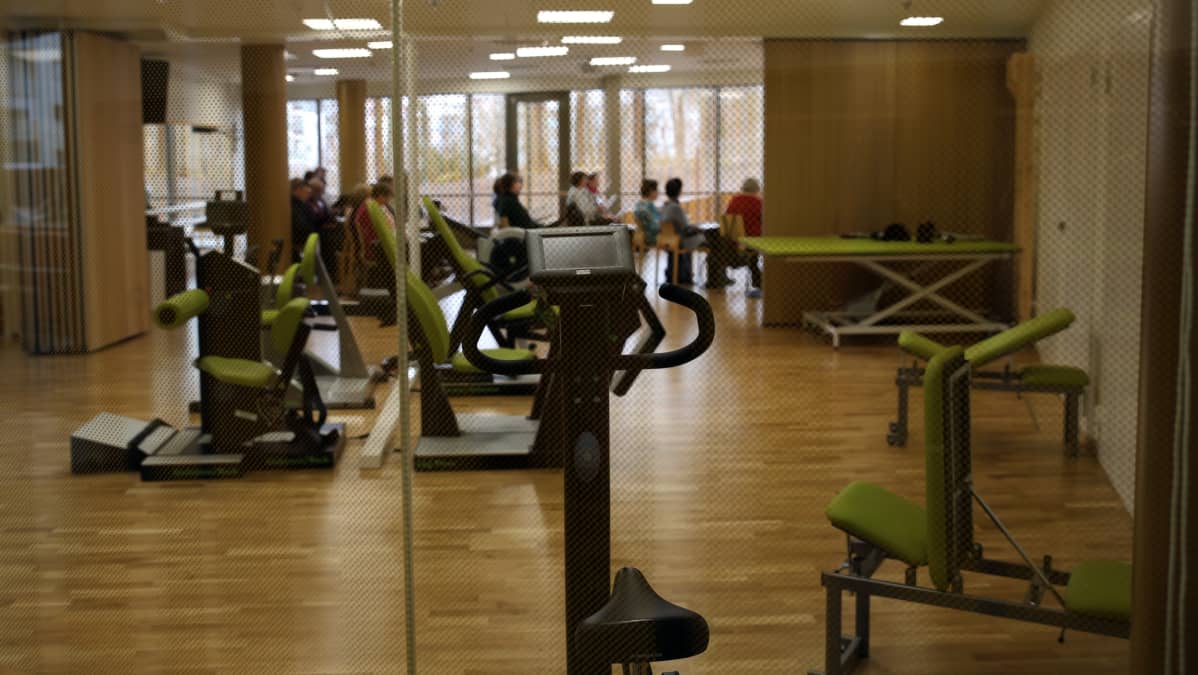 Kuvassa kuntosali, jonka takaosassa ryhmä ikäihmisiä keppijumppaa.