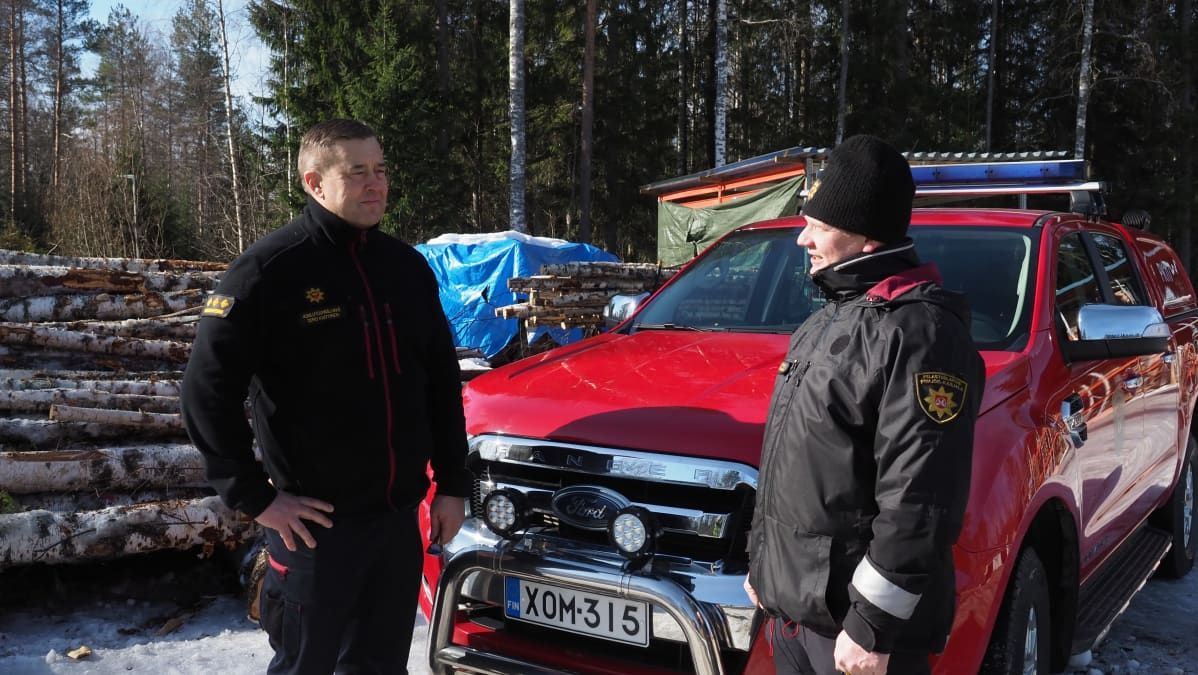 Kaksi pelastuslaitoksen työntekijää punaisen auton edessä ulkona.