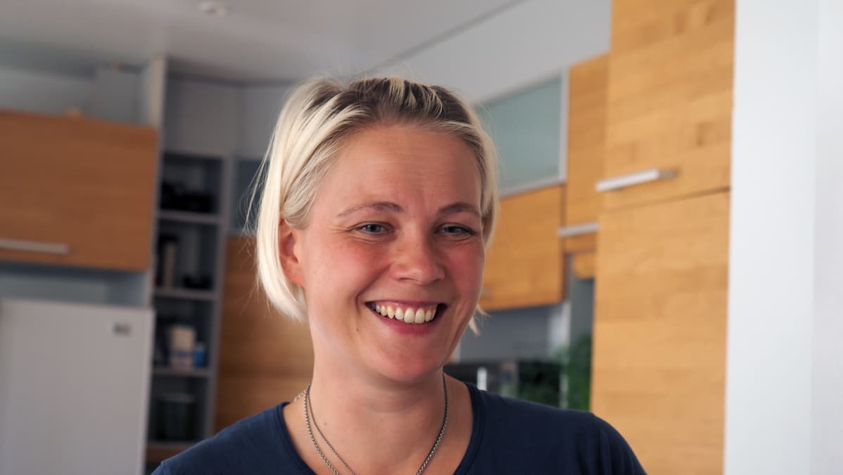 Nainen hymyilee, keittiö näkyy taustalla