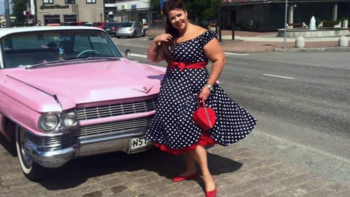 Nainen pilkullisessa mekossa poseeraa vaaleanpunainen Cadillacin vierellä.