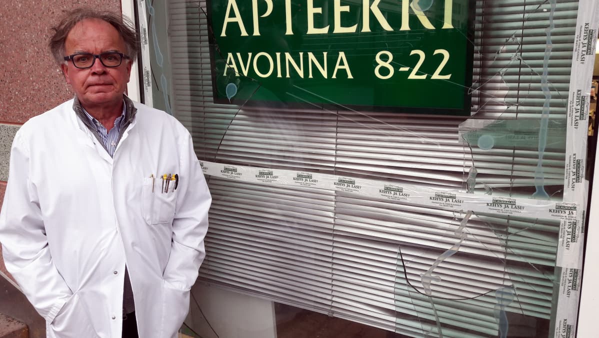 Juha Nuutinen seisoo valkoisessa takissa hajonneen apteekin ikkunan vieressä.