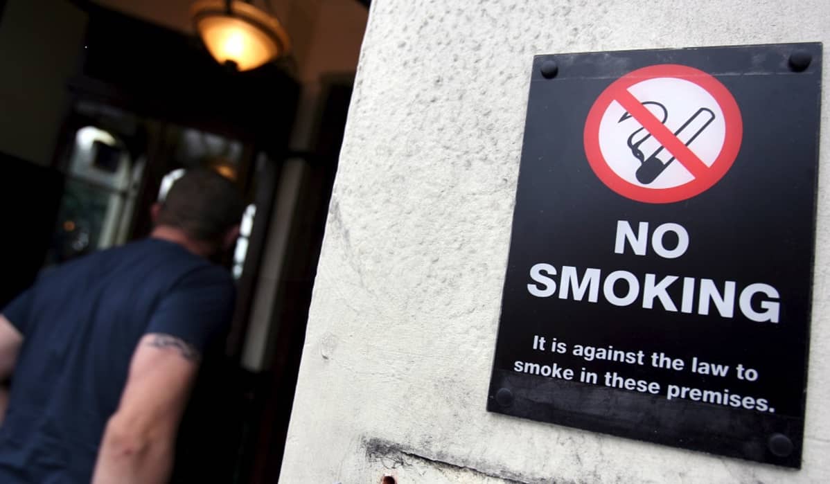 Kuvassa kyltti, joka kertoo tupakointikiellosta. Kyltissä lukee No smoking. Taustalla mies kulkee kulman ohi. Kuva on otettu Lontoossa.
