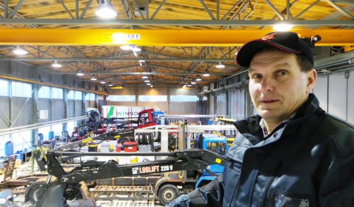 mies seisoo hallissa jossa on metsäkoneita ja kuorma-autoja