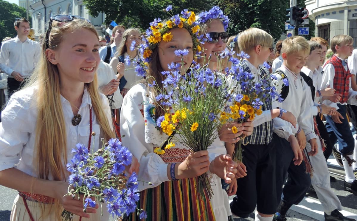 Hymyileviä kansallispukuisia tyttöjä, joilla on käsissään kukkia.